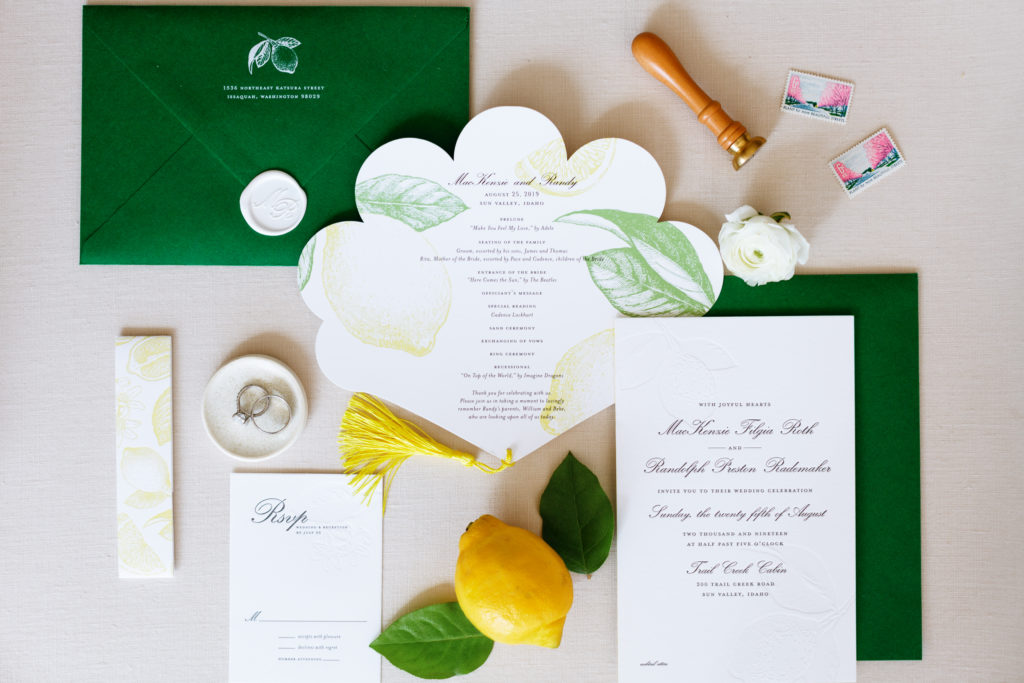A flat lay of lemon-themed wedding invitations, green envelopes, and the bride's wedding rings.