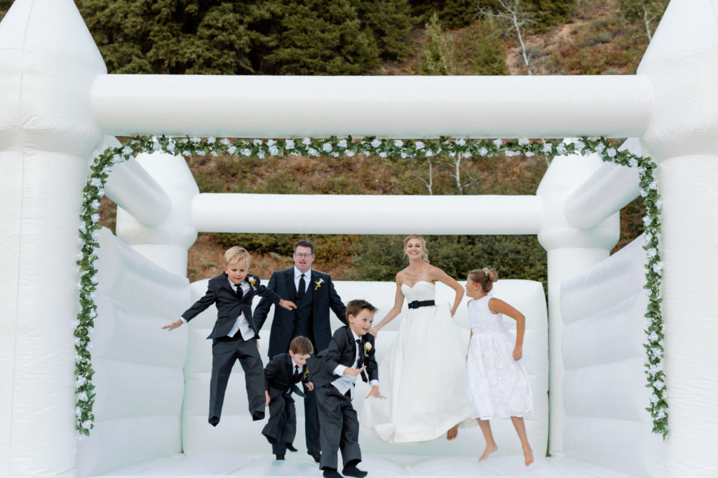 Bride, groom, and four kids jump together in a white bouncy house.