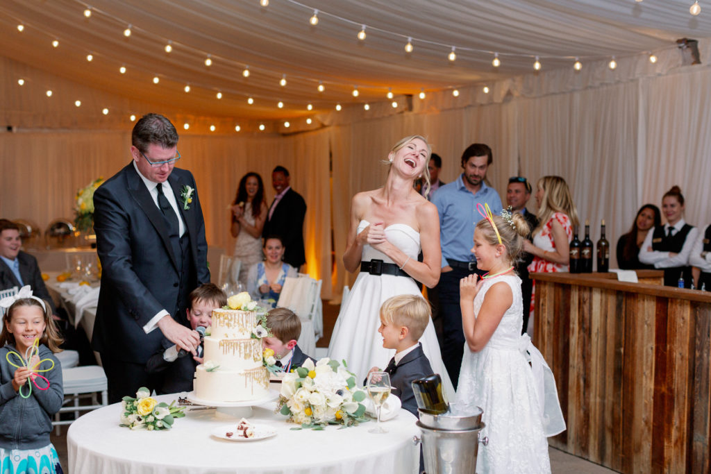 Bride laughs as her groom cuts the cake and children look on.
