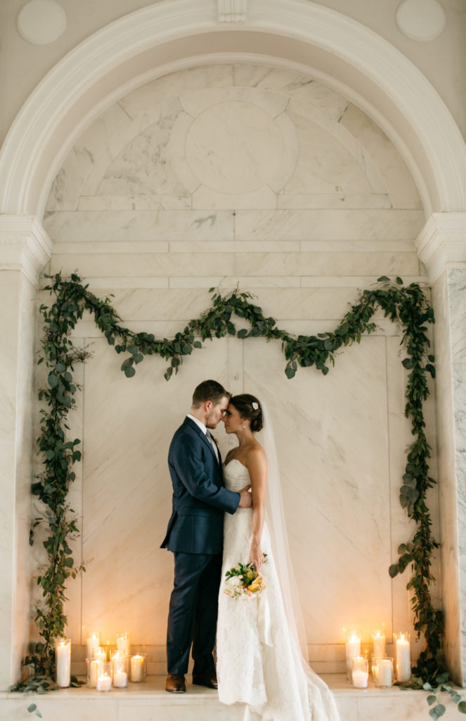 A bride and groom bump foreheads in front of a white marble wall decorated with greenery and white candles. 