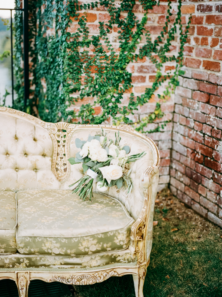 A white and green bridal bouquet rests on a green vintage couch in front of a brick wall at an outdoor wedding venue near Atlanta.